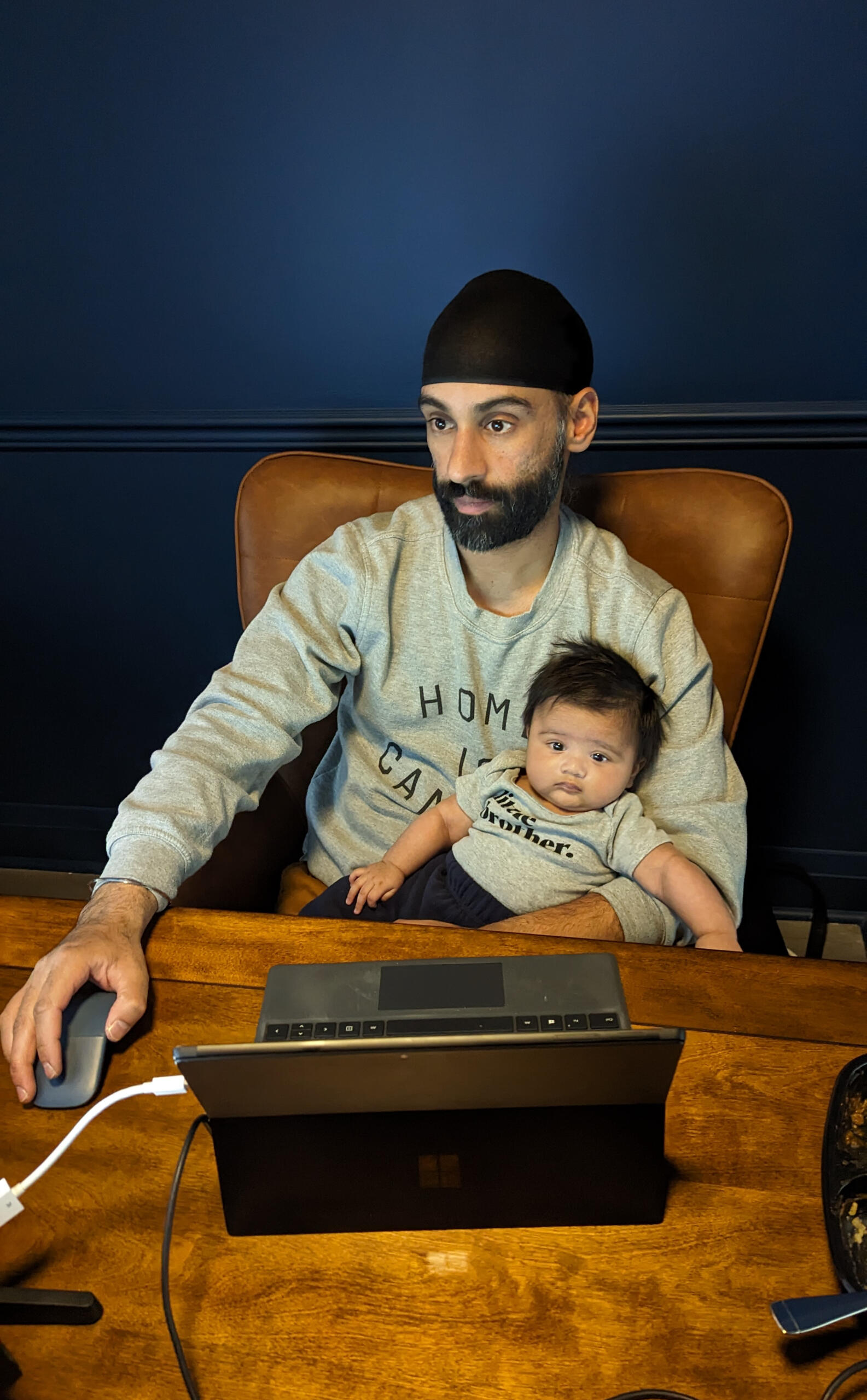 Manny Bahia holding his second newborn, sitting at his home office desk.